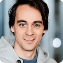 Smiling man with dark hair and blue eyes wearing a light gray hoodie.